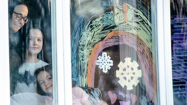 À la fenêtre d'une maison, une maman avec ses deux filles sourient. Un arc-en-ciel est dessiné dans la fenêtre.