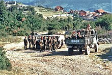 Three trucks of soldiers idle on a country road in front of trees and red roofed houses. The rear truck has KFOR painted on is back.