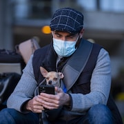 Un homme portant un masque regarde son cellulaire en compagnie de son petit chien. 