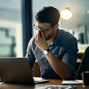 Un homme se tient le nez en signe d'épuisement devant son ordinateur portable posé sur une table. 