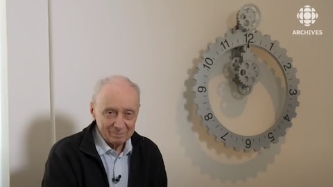 Le cinéaste et réalisateur Fernand Dansereau assis à côté d'une horloge.