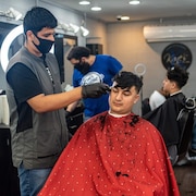 Des hommes sont assis dans des chaises et se font couper les cheveux par des barbiers qui portent des masques médicaux.