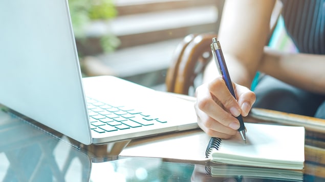 Une main de femme prend des notes dans un cahier, à côté d'un ordinateur portable. 