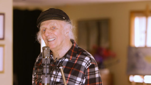 Gilles Vigneault, qui porte une chemise à carreaux et une casquette noire, sourit devant un micro.