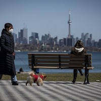 Une femme avec un masque passe à distance d'un banc public sur lequel une autre femme est assise. Cette dernière regarde la ville de Toronto.