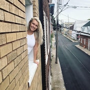 Marylene Levesque a le haut du corps sorti d'une fenêtre, et la vue derrière elle est une rue de la basse-ville de Québec. Elle a les cheveux blonds et est souriante.