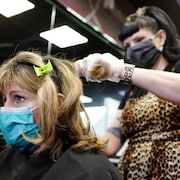 Une coiffeuse est debout derrière une cliente assise. Les deux portent un masque sur leur visage.
