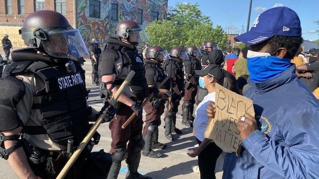 Des policiers antiémeute forment une ligne devant des manifestants placés devant eux. Un homme tient une pancarte où on peut lire : «Black lives matter».