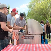 Quatre hommes s'activent autour du barbecue avec en arrière-plan quatre femmes qui discutent dans le parc.