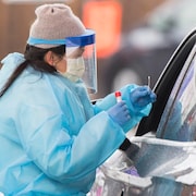 Une femme portant des vêtements et une visière de protection recueille un échantillon de salive d'un automobiliste aux fins d'un test de dépistage de la COVID-19.