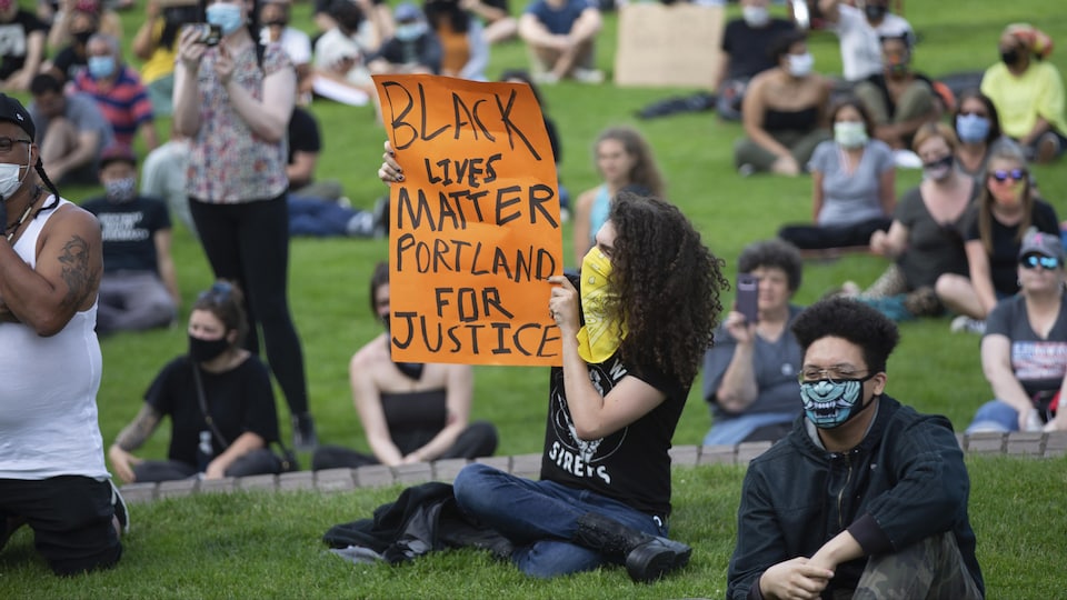 Des manifestants assis sur la pelouse dans un parc, une femme tient une affiche sur laquel il est écrit «Black lives matter, Portland for justice».