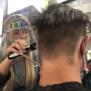 Une coiffeuse, portant un équipement de protection contre le coronavirus, rase les cheveux d'un client.
