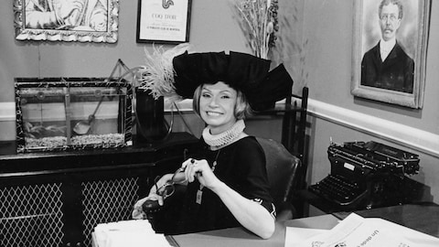 Monique Mercure, dans un costume des années 1920, est assise à un bureau, souriante. 