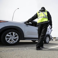 Un agent vêtu d'une veste jaune et avec des documents à la main s'approche d'une automobile.