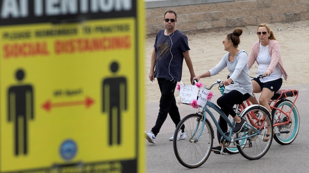 Deux femmes font du vélo côte à côte, dépassant un piéton, près d'un panneau prônant la distanciation sociale.