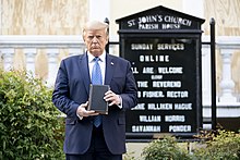 President Donald Trump, dressed in a dark blue suit with a light blue tie and white dress shirt, holds a copy of the Bible in front of Ashburton House, a former private residence which now serves as the priory house of St. John's Episcopal Church just north of Lafayette Square. St. John's is popularly known as the "Church of the Presidents" because every president since James Madison has attended services there at least once, typically on the day of their inauguration.