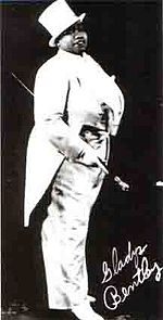 A publicity photo of a stout African American woman in white tuxedo with tails and top hat, carrying a cane and her signature in the lower right corner.