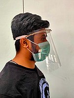 Person wearing a face shield over a green surgical mask. A simple 3D-printed face shield: curved visor, drawstring lanyard, sheet of transparent plastic curved from side to side.