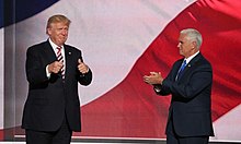 Donald Trump and his running mate for vice president, Mike Pence. They appear to be standing in front of a huge screen with the colors of the American flag displayed on it. Trump is at left, facing toward the viewer and making "thumbs-up" gestures. Pence is at right, facing Trump and clapping.