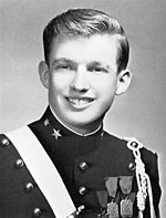 A black-and-white photograph of Donald Trump as a teenager, smiling and wearing a dark pseudo-military uniform with various badges and a light-colored stripe crossing his right shoulder