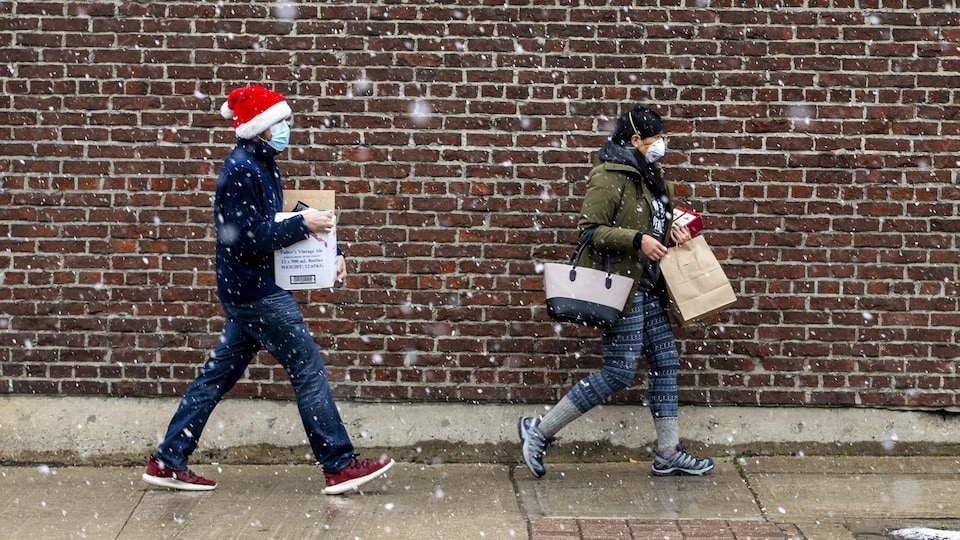 Deux personnes marchent sous la neige avec des sacs de magasinage à Toronto le 22 novembre 2020. L'une d'elles est coiffée d'une tuque de Noël.