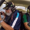 Un homme avec une barbe blanche tient un téléphone intelligent à bord d'un petit avion avec un pilote à ses côtés, en plein vol. 
