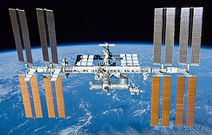 A foreward view of the International Space Station backdropped by the limb of the Earth. In view are the station's four large, maroon-coloured solar array wings, two on either side of the station, mounted to a central truss structure. Further along the truss are six large, white radiators, three next to each pair of arrays. In between the solar arrays and radiators is a cluster of pressurised modules arranged in an elongated T shape, also attached to the truss. A set of blue solar arrays are mounted to the module at the aft end of the cluster.