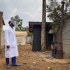 Un employé médical, vêtu d'un sarreau et d'une visière, se tient sur un terrain vague en République démocratique du Congo. 