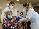 Elderly woman roles up sleeve as two nurses administer a vaccine