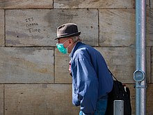Un homme, avec chapeau, veste bleu, masque lui couvrant la bouche et le nez. Derrière sur un mur un grafiti : "It's Corona time " (C'est le temps du Corona).