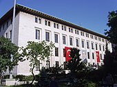 A four-story, white flat-roofed building with two Turkish flags and a portrait on the exterior