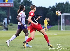photo of Quinn in red Washington Spirit kit (right) with soccer ball in mid-air after being cleared to defend against Orlando Pride forward Chioma Ubogagu wearing purple kit