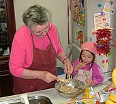 Une grand-mère portant un tablier bordeaux sur une blouse rose, racle au-dessus du plat en inox de pâte qu'elle vient de confectionner l'un des fouets de son mixeur-batteur tandis qu'à côté d'elle sa petite-fille habillée de rose fuchsia et portant un tablier identique mais rose pâle, se lèche le doigt utilisé pour gouter la pâte prélevée sur le second fouet.