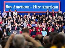 Trump speaks at a podium, with a crowd in front of and behind him. A banner behind him reads "Buy American – Hire American"