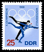 A postage stamp with a blue background and two figure skaters skating, the date 1968 is centred on the top of the stamp along with the Olympic rings. The word "Winterspiele" is written down the right side, the words "X Olympische" are written down the left side. The number 25 is in the lower-left corner and the letters "DDR" are in the lower right corner