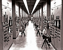 A long corridor with many consoles with dials and switches, attended by women seated on high stools