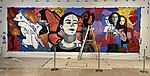 Angennette Escobar stands on a ladder while painting a mural, Metamorphosis (triptych), in the Portland Art Museum. The mural is a collaboration between artists Hector H Hernandez, Christian Barrios, and Angennette Escobar. The mural depicts Frida Kahlo at three different ages, addressing three different aspects of her identity. One as a mestiza, another as a non-binary or gender fluid person, and one addresses her health condition.