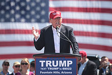 Trump speaking in front of an American flag behind a podium, wearing a black suit and red hat. The podium sports a blue "TRUMP" sign.