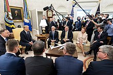 Donald Trump and Andrzej Duda sit in a packed Oval Office, surrounded by other officials and members of the press. Only the press are masked.