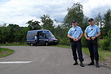 At the side of a road, in the foreground, French gendarmes and, in the background, two more with a gendarmerie van.