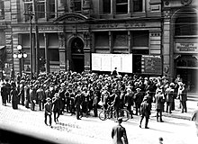 Black and white image of building exterior, including a group of people reading news boards