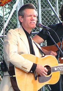 Singer Randy Travis, wearing a tan suit and singing into a microphone while playing an acoustic guitar.