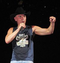 Country singer Kenny Chesney, singing with one arm extended and the other holding a microphone.