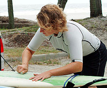 Photo of a woman bent over surfboard rubbing bar of solid wax against the board with palm trees and ocean in background