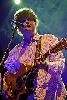 Ron Sexsmith wearing a striped shirt, playing an acoustic guitar and singing into a microphone onstage