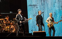 U2 performing on a concert stage. The Edge and Adam Clayton, playing guitars, flank Bono in the foreground, while Larry Mullen, Jr. is behind a drum kit in the background on the left side.