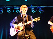 Country music singer Rodney Crowell, seated on a stool and playing an acoustic guitar