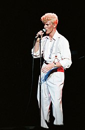 A man with blonde hair and a white suit holding a microphone