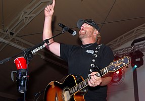 Singer Toby Keith onstage, smiling and holding a guitar behind a microphone while pointing upward