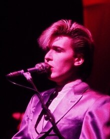 Singer with quiff upstanding on stage with microphone on stand and atmospheric deep red lighting